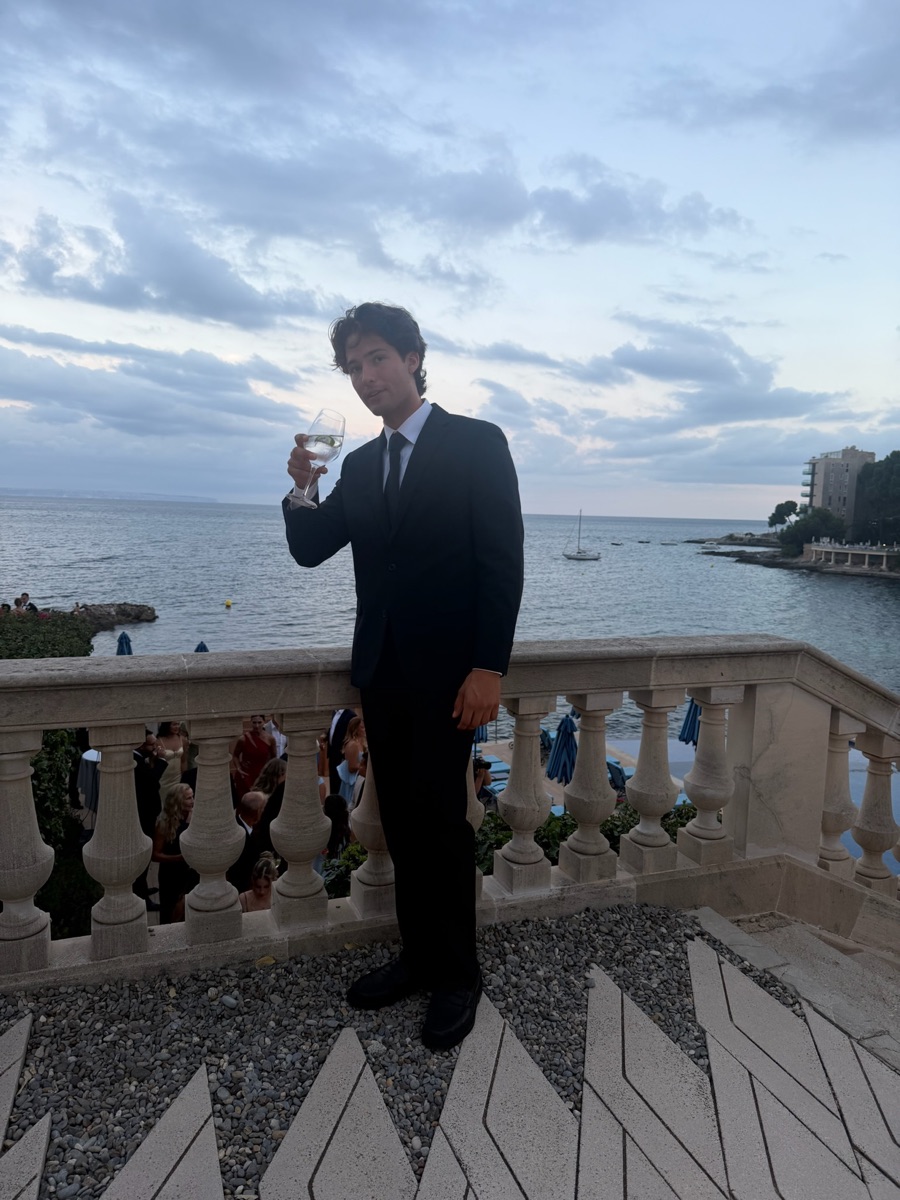 Braiden in a black suit raising a glass on a seaside terrace at sunset